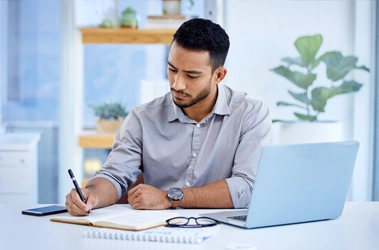 A man sits at a table, focused on his laptop, with a neutral background.